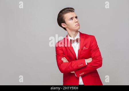 Need to think. Portrait of confused thoughtful gentleman in red tuxedo and bow tie crossing hands and looking up with doubtful face, pondering idea. indoor studio shot isolated on gray background Stock Photo