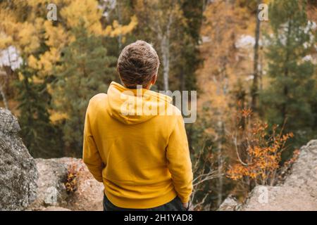 A man in a yellow hoodie stands on top of a mountain, admiring the magnificent landscape of the yellow autumn forest from above. Selective focus.