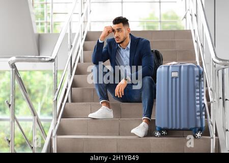 Flight Cancellation. Upset Arab Man Sitting With Luggage At Stairs In Airport Stock Photo