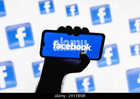 Smartphone with the Facebook social network logo on the screen in a clenched hand on the background of Facebook logos Stock Photo