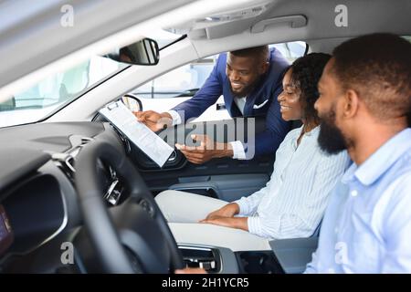 Young Black Couple Sitting Inside New Car In Showroom, Listening Professional Salesman Stock Photo