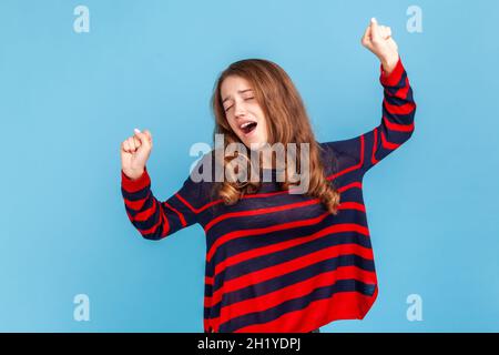 Portrait of sleepless woman wearing striped casual style sweater, yawning and raising hands up, feeling fatigued, standing with close eyes. Indoor studio shot isolated on blue background. Stock Photo