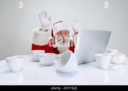 Cropped portrait of senior man wearing Santa Claus costume sitting at the desk full of empty coffee cups and looking on laptop Stock Photo