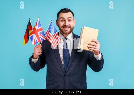 Satisfied businessman or teacher wearing official style suit, holding flags of Germany, Great Britain and USA and book, looking at camera with smile. Indoor studio shot isolated on blue background. Stock Photo