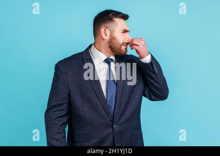 Bad smell. Portrait of bearded man in suit holding breath and grimacing in disgust, expressing repulsion to stink, fart gases, intolerable odor. Indoor studio shot isolated on blue background. Stock Photo