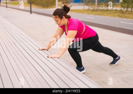 High-angle view of motivated fat young woman training doing push ups using street bench in city park at summer morning. Stock Photo