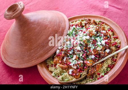 Chicken cous cous tagine with pomegranate seeds, feta cheese and herbs in a terracotta tagine with a serving spoon on a red tablecloth Stock Photo