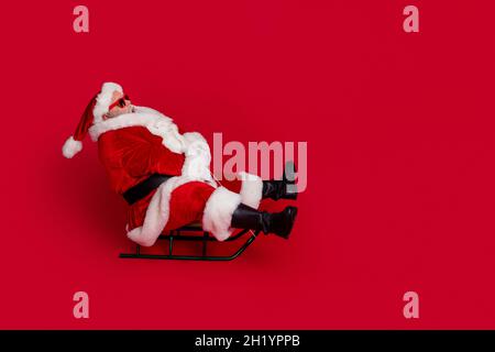 Full size profile photo of funny santa go by sled wear cap glasses coat pants shoes isolated on red color background Stock Photo