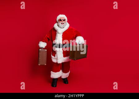 Photo of funny excited santa claus wear red headwear hat dark glasses smiling hurrying check in isolated red color background Stock Photo