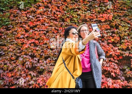 Westminster, London, UK. 19th Oct, 2021. Two women take selfies with the vibrant wall. People walk past the vibrant wall. The virginia creeper (Parthenocissus quinquefolia) at the Admiralty Citadel near Horseguards Parade is slowly turning to its vibrant red and orange autumn colours, on a very mild day in London with temperatures around 19 degrees. Credit: Imageplotter/Alamy Live News Stock Photo