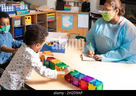 Education Preschool 3-4 year olds female teacher taking notes as two boys play together with magnet tiles, all wearing face masks to protect against C Stock Photo