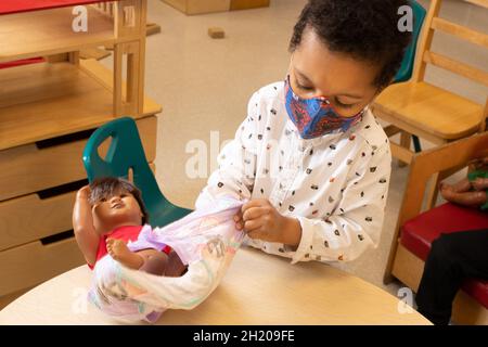 Education Preschool 3-4 year olds boy dressing doll in family area, wearing face mask to protect against Covid-19 infection Stock Photo