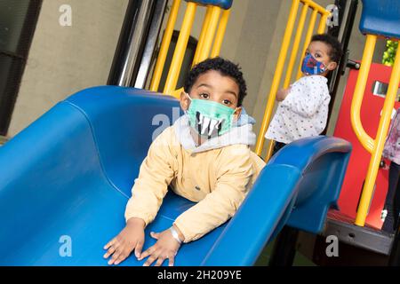 Education Preschool playground boy playing on slide wearing face mask to protect against Covid-19 Stock Photo