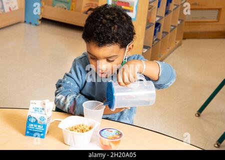 Education Preschool 3-4 year olds boy pouring water at breakfast, ceeral, milk and fruit on table before him Stock Photo