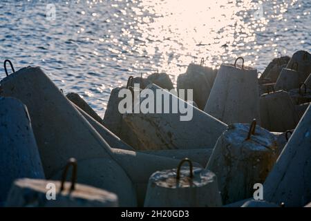 Large concrete breakwaters tetrapods near blue sea with glare of sunlight on water surface, macro. Blocks and stones for protection coastline from sto Stock Photo