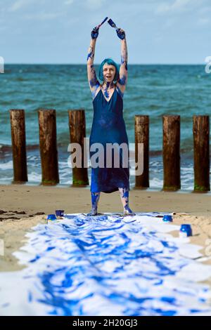 Young woman performance artist in dark blue dress smeared with blue gouache paints happily dancing hands up on beach. Canvas for expressive painting w Stock Photo