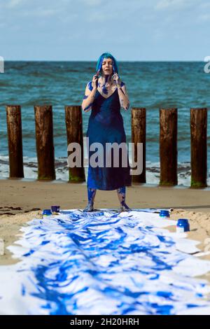 Young woman performance artist in dark blue dress smeared with blue gouache paints happily dancing hands up on beach. Canvas for expressive painting w Stock Photo