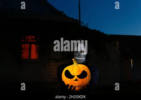 Grim reaper holding halloween pumpkin head. Man in death mask with fire flame in eyes on night sky, light window background. Halloween holiday concept Stock Photo