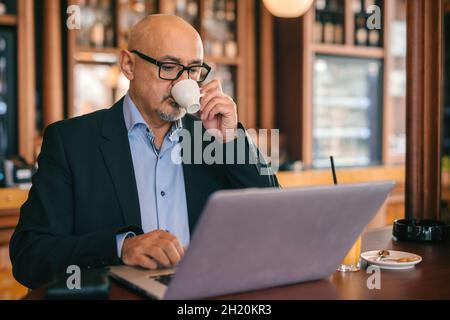 Bearded senior businessman in suit using laptop and drinking coffee while sitting in cafeteria. Stock Photo
