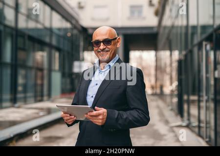 Smiling bearded senior in suit holding tablet while standing outdoors. Stock Photo