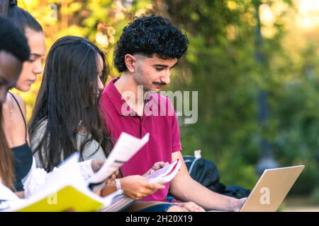 Diverse young people studying with laptop and papers sitting on a bench outdoors Stock Photo