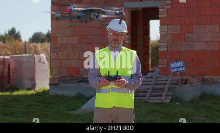 Drone operator holding remote controller controls an aircraft in front of construction house site Stock Photo