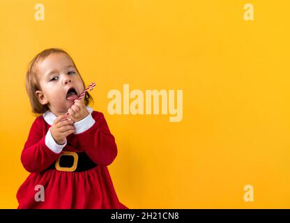 Banner Portraite Cute Cheerful Chubby Baby Girl in Santa Suite Holding Eating Caramel Candy At Yellow Background. Child Play Christmas Celebrating Stock Photo