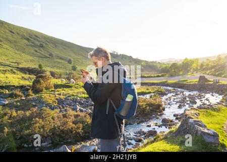 Side view young explorer man trekking in mountains, using mobile phone for GPS application. Traveler man texting messages and browsing on Internet via Stock Photo