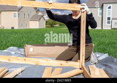 a woman is building a wooden fence or keep on tarp in her backyard. A concept for wood work. Furniture installation DIY project. She wears gloves to p Stock Photo