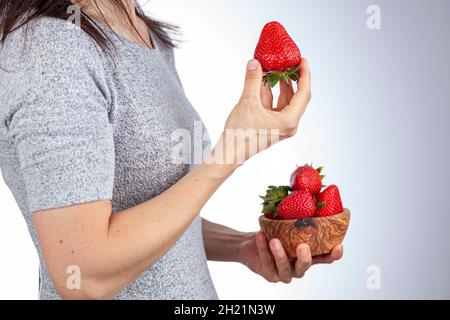 Caucasian young woman wearing short sleeve t-shirt is eating strawberry snack. Side view photo shows her holding a wooden bowl of berries. Summer, hea Stock Photo