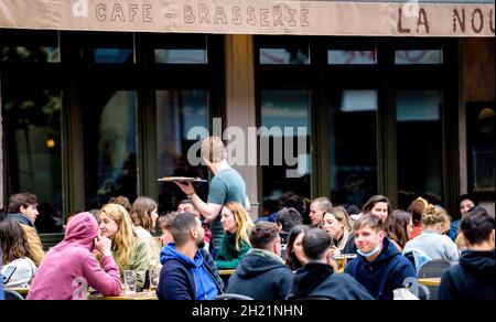 Waiter with tarte flambee food walking between tables of Nouvelle poste terrace full with people after covid-19 outbreak Stock Photo