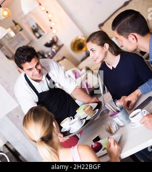 Polite friendly waiter bringing ordered dishes to friends in tearoom Stock Photo