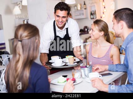 positive waiter bringing ordered dishes to friends in tearoom Stock Photo