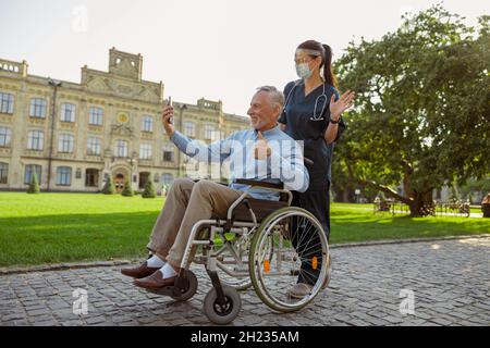 Mature man, recovering patient in wheelchair making a video call using smartphone on a walk with nurse wearing face shield and mask Stock Photo