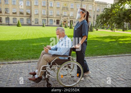 Side view of senior man recovering patient in wheelchair using smartphone on a walk with nurse wearing face shield and mask Stock Photo
