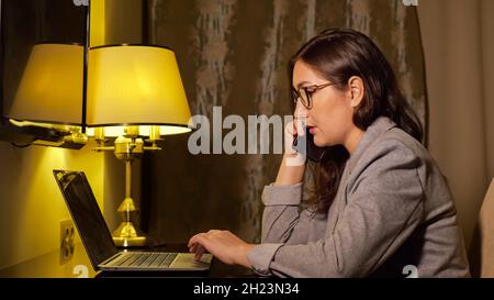 woman in a business suit in the evening works at a laptop and talks on the phone Stock Photo