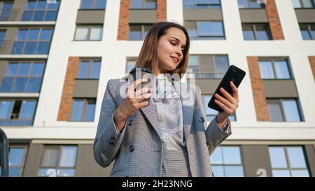 woman in a business suit looks at the phone, while holding a plastic cup on the background of a building Stock Photo