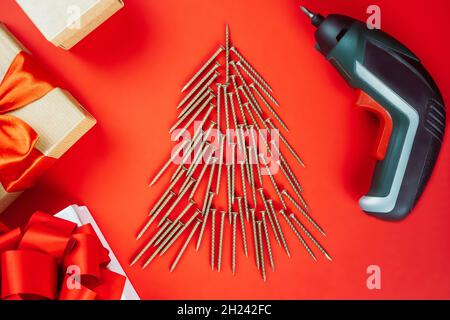 Long golden self-tapping screws, laid out in shape of Christmas tree, screwdriver and gift boxes on red background Stock Photo