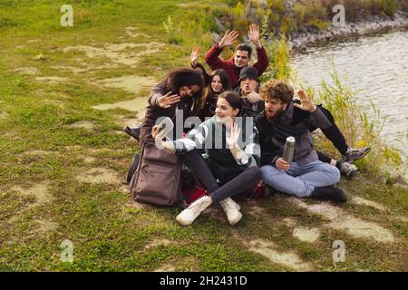 Full-length portrait of group of friends having nice picnic, taking selfie near lake, river on warm autumn day. Countryside hiking Stock Photo