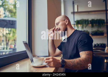 Serious hipster millennial man works at laptop while sits in cafe with coffee. Freelance concept Stock Photo