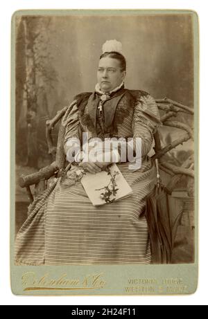Victorian cabinet card of wealthy prosperous middle class, mature religious Christian woman, looking serious, stern, probably Catholic,  wearing an elaborate dress with leg of mutton sleeves, lace cap, crucifix on display showing she is devout, an umbrella by her side, holding a newspaper to show she is educated, from the studio of Debenham & Co., Victorian Lodge, Western-Super-Mare, Somerset, England, U.K. dated June 1894