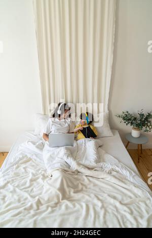 Mom and Son work and study lying together in bed using laptop computer and digital tablet in bedroom Stock Photo
