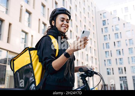 Beautiful young woman working as a courier with a delivery bag on her back, holding mobile phone and smiling Stock Photo