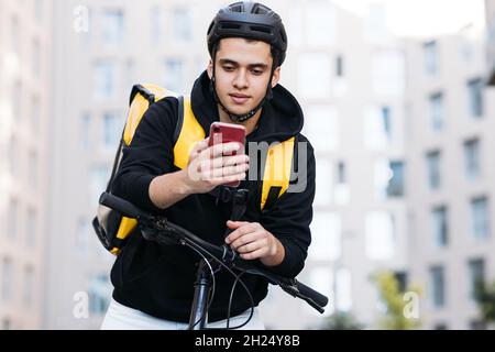 Male courier leaning on the handlebar of a bicycle wearing food delivery backpack and cycling helmet looking at mobile phone Stock Photo