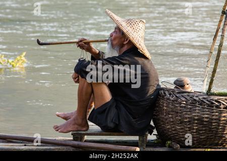 Elderly traditional cormorant fisherman smokes a wooden pipe on his raft on the Li River, Xingping, China. Stock Photo