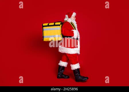 Photo of funny cheerful man pensioner dressed santa claus costume walking carrying delivery rucksack smiling isolated red color background Stock Photo
