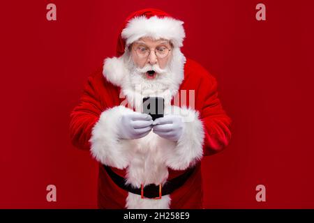 Photo of funky impressed man pensioner dressed santa claus costume chatting modern device big eyes isolated red color background Stock Photo