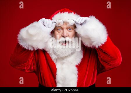 Photo of amazed surprised retired man wear santa claus costume arms spectacles open mouth isolated red color background Stock Photo