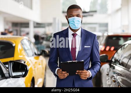 Young Black Car Salesman Wearing Medical Mask Posing At Workplace In Showroom Stock Photo