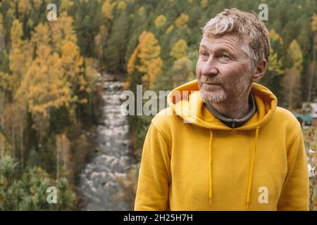 An authentic portrait of a handsome unshaven adult male traveler in a yellow hoodie on top of a mountain, against the background of an autumn forest. Stock Photo